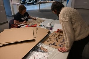 Two people at opposite ends of a table are writing on large cardboard signs